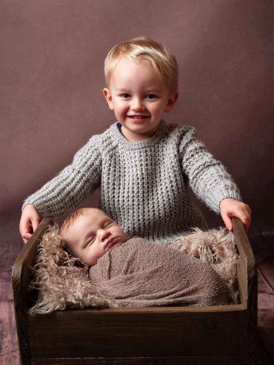 Smiling toddler behind a wooden cot holding a sleeping newborn
