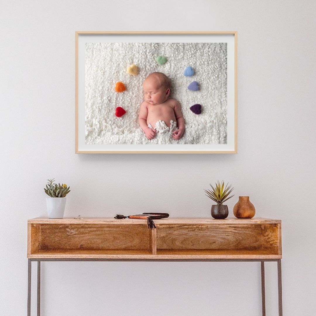 Framed newborn photo on a wall above a wooden console table, showing a sleeping baby surrounded by colourful felt hearts on a white blanket